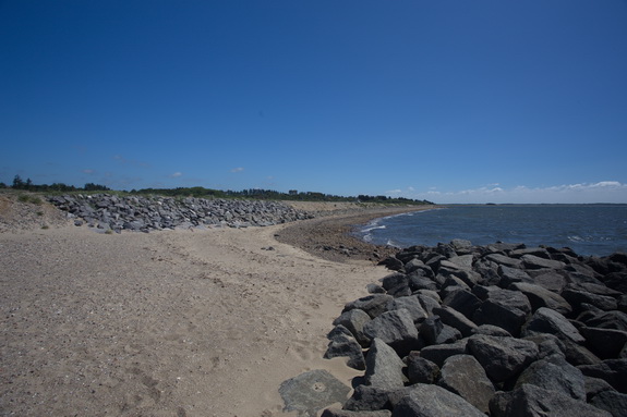 Øst for bølgebryderne mangler sandet, som skulle have været med til at yde en naturlig beskyttelse af kysten. Værst er det lige efter bølgebryderne, hvor kysten er udsat for fænomenet læside-erosion. Her gnaver fjorden ind i en dyb bue. Som modtræk har kystbeskyttelseslaget måttet beskytte skråningen på dette sted med granitblokke. (Foto: Peter Jessen)
